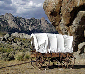 A white canvas wagon sits in front of a large rock buttress with mountains in the distance.