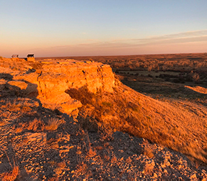 Warm light from the setting sun gives an orange glow to a rock bluff overlooking a grassland.